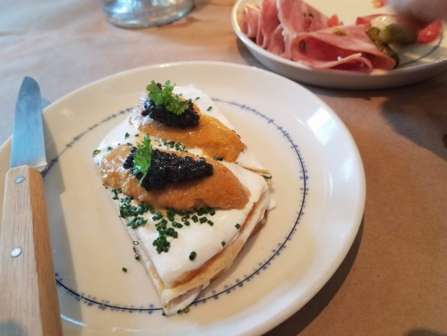 A plate with two crepes topped with cream, caviar, and herbs, alongside a dish of sliced meats.