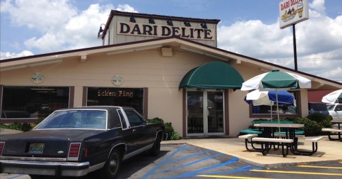 Exterior of Dari Delite restaurant with a black car parked outside and picnic tables under umbrellas. Bright blue sky above.