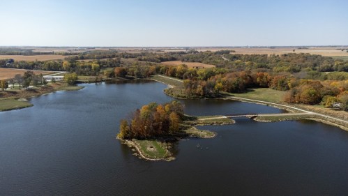 Aerial view of a serene lake surrounded by autumn-colored trees and fields under a clear blue sky.