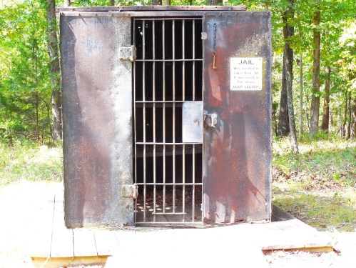 A weathered metal jail cell with a barred door, set in a wooded area, featuring a sign that reads "JAIL."