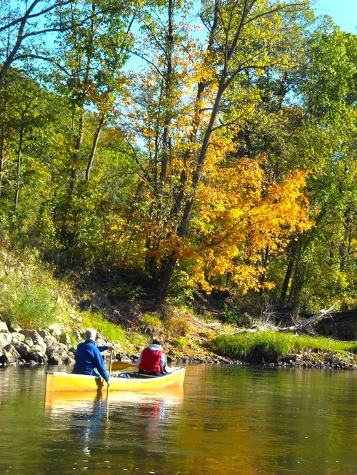 Two people in a canoe paddle along a calm river surrounded by trees with vibrant autumn foliage.