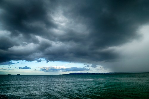 Dark, stormy clouds loom over a calm sea, with a hint of blue sky peeking through the clouds on the horizon.