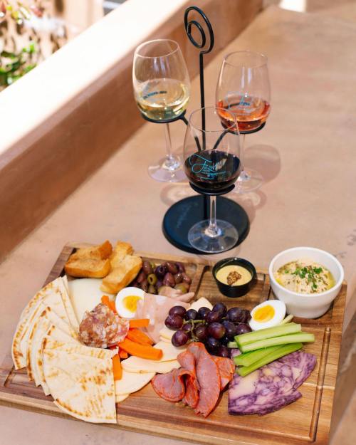 A wooden platter with assorted cheeses, meats, vegetables, and grapes, accompanied by three glasses of wine.
