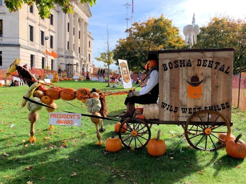 A festive display featuring a pumpkin-drawn cart with a scarecrow, surrounded by autumn decorations and pumpkins.