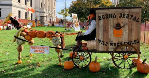 A festive display featuring a pumpkin-drawn cart with a scarecrow, surrounded by autumn decorations and pumpkins.