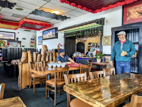 A cozy restaurant interior with wooden tables, a bar area, and two men interacting near the entrance.