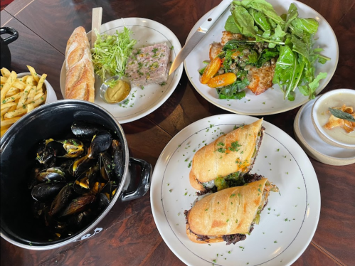 A table spread featuring mussels, a salad, a meat pâté, fries, and two sandwiches, garnished with herbs.