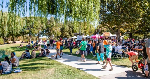 A lively park scene with people walking, sitting, and enjoying a festival under trees and tents.