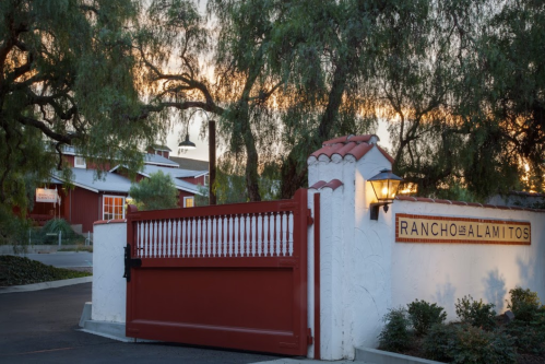 Entrance gate of Rancho Los Alamitos, featuring a red gate and rustic sign, surrounded by trees at sunset.