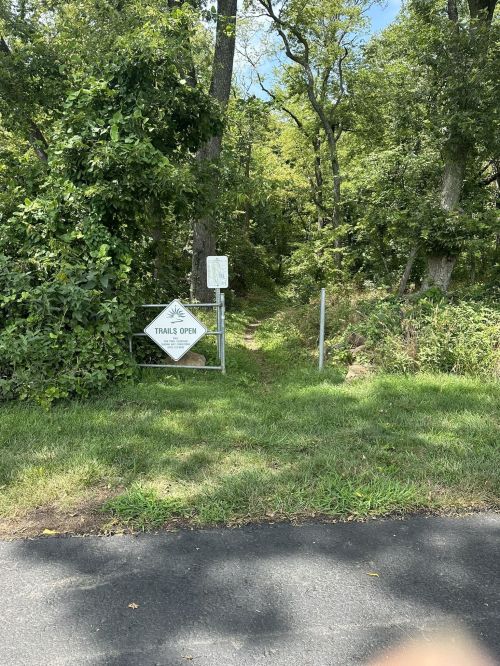 A sign reading "Trail's Open" marks the entrance to a wooded path surrounded by greenery.