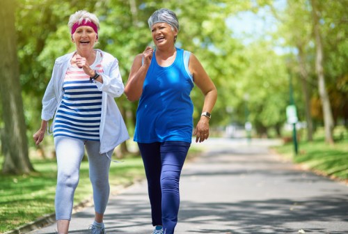 Two older women jog together in a sunny park, wearing athletic clothing and smiling joyfully.