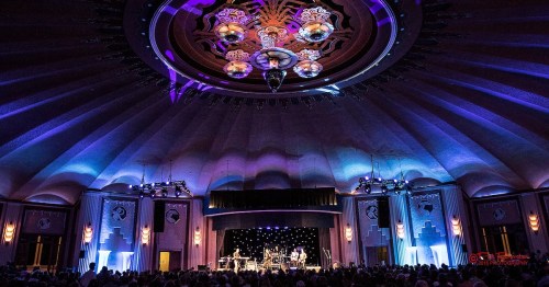 A vibrant concert scene in a grand venue with colorful lighting and a decorative ceiling, filled with an audience.