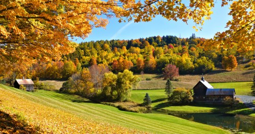 A picturesque autumn landscape with colorful trees, a serene pond, and rustic buildings under a clear blue sky.
