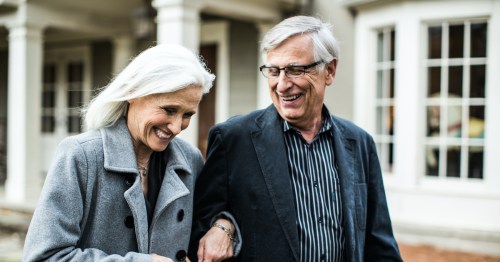 A smiling elderly couple walks hand in hand outside a house, enjoying each other's company.
