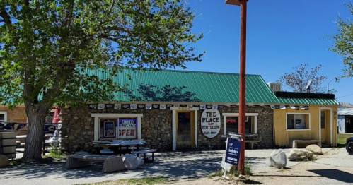 A rustic stone building with a green metal roof, featuring signs that say "The Place" and "Open," surrounded by trees.