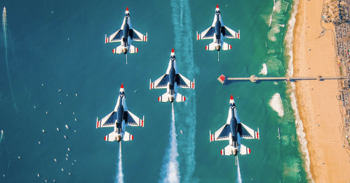 Five fighter jets flying in formation over a beach and pier, leaving trails of smoke against a clear blue sky.