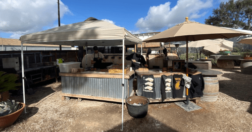 Outdoor food stall with tents, serving area, and tables surrounded by plants and a cloudy sky.