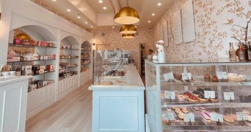 A bright bakery interior with shelves of treats, a glass display case of pastries, and stylish golden pendant lights.