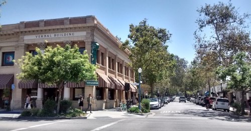 A sunny street scene featuring the Verbal Building, trees, and people walking along a charming downtown area.