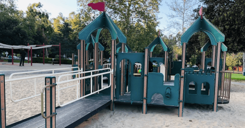 A colorful playground structure with slides and climbing areas, surrounded by trees and sandy ground.