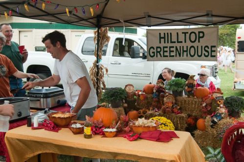 A vendor at a market booth with pumpkins and decorations, serving food under a tent labeled "Hilltop Greenhouse."