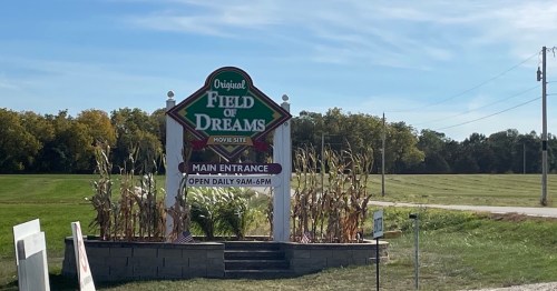 Sign for the Original Field of Dreams, featuring the main entrance and hours of operation, set against a green field.