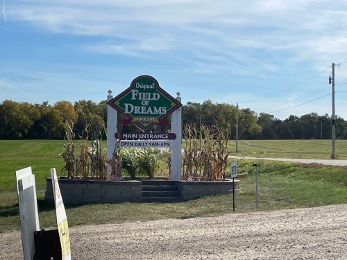 Sign for the Field of Dreams, featuring "Main Entrance" and hours, surrounded by cornfields under a clear blue sky.