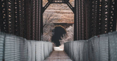 A perspective shot looking down a fenced walkway of an old bridge towards a dark tunnel entrance in the distance.