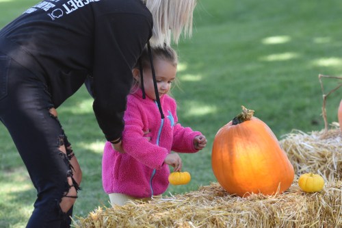 A woman helps a young girl pick up a small pumpkin next to a large pumpkin on a hay bale in a grassy area.