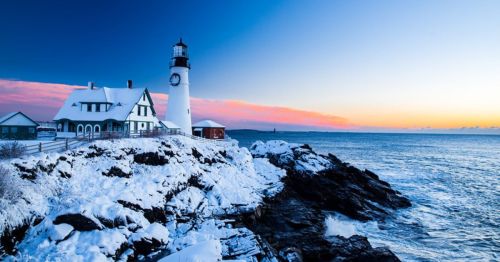 A snowy lighthouse by the ocean at sunset, with rocky shores and a colorful sky in the background.