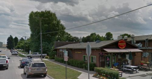 A cozy donut shop with a wooden exterior, surrounded by greenery and parked cars on a cloudy day.