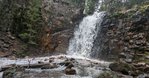 A cascading waterfall flows over rocky cliffs, surrounded by lush greenery and splashing into a river below.