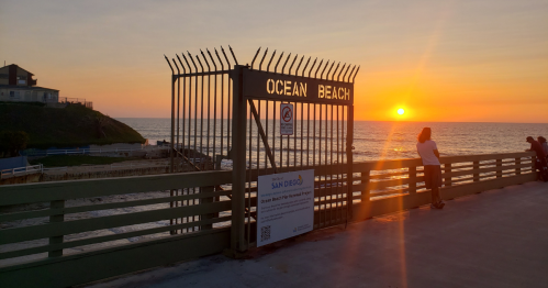 A sunset over the ocean viewed through a gate labeled "Ocean Beach," with people enjoying the scene nearby.