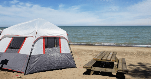 A tent on a sandy beach with a picnic table nearby, overlooking a calm lake under a blue sky.