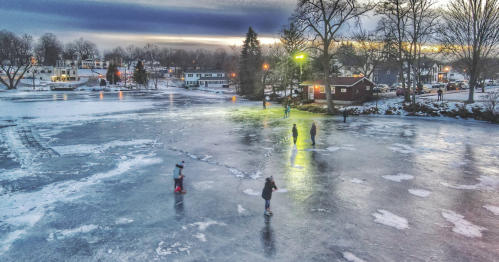People ice skating on a frozen lake at sunset, surrounded by snow-covered trees and houses.