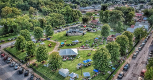 Aerial view of a park with a stage, tents, and people enjoying a community event surrounded by trees and parked cars.