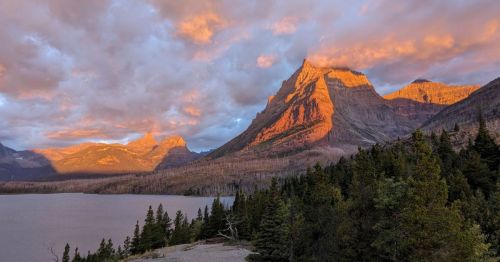 Sunset over rugged mountains reflecting warm hues, with a serene lake and lush trees in the foreground.