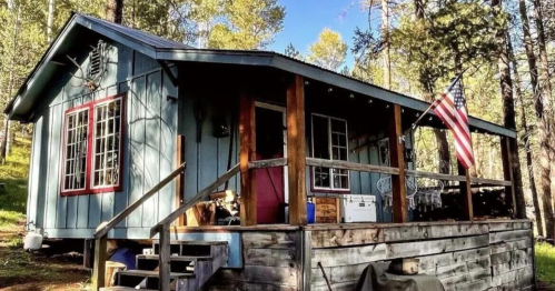 A cozy blue cabin with a porch, surrounded by trees, featuring an American flag and rustic wooden steps.