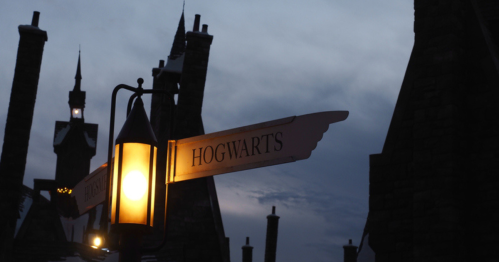 A lantern points towards a sign reading "Hogwarts" against a twilight sky, with castle-like structures in the background.