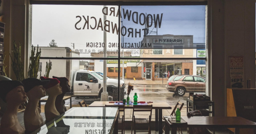 View from inside a shop with a sign reading "Woodward Throwbacks," showcasing a rainy street and parked cars outside.