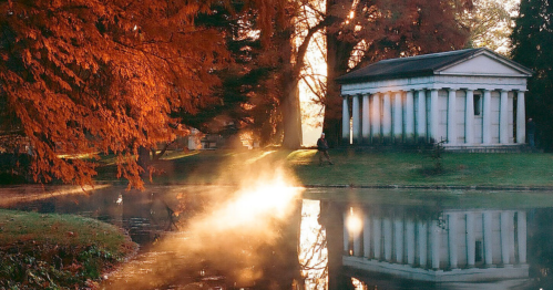 A serene landscape featuring a pond, autumn trees, and a classical building reflecting in the water at sunrise.