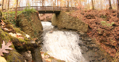 A serene waterfall flows beneath a wooden bridge, surrounded by autumn foliage and rocky terrain.