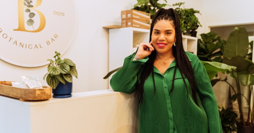 A woman in a green shirt stands by a plant-filled counter at a botanical bar, smiling confidently.
