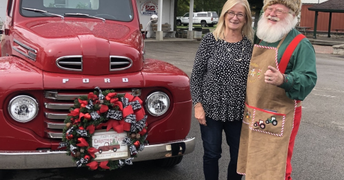 A woman stands next to a smiling Santa in an apron, beside a red vintage Ford truck decorated for Christmas.