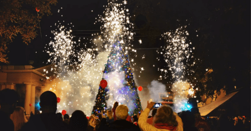 A crowd gathers around a brightly lit Christmas tree, surrounded by fireworks and festive decorations at night.
