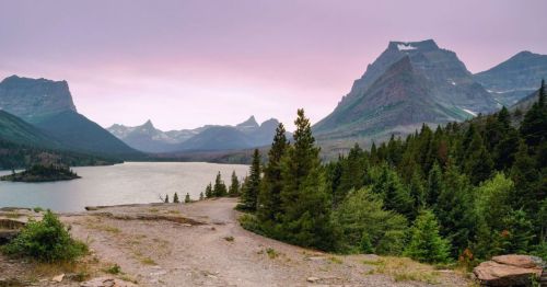 Scenic view of a lake surrounded by mountains and forests under a pinkish sky at dusk.