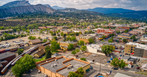 Aerial view of a town surrounded by mountains, featuring buildings, trees, and a railway in the foreground.