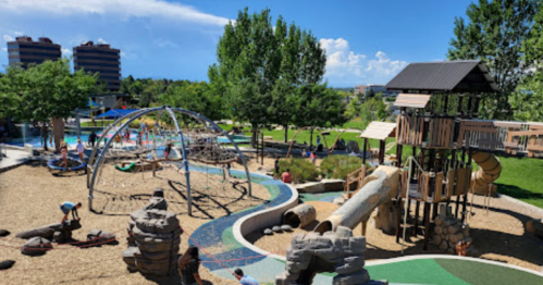 A vibrant playground with climbing structures, slides, and a water area, surrounded by green trees and blue skies.