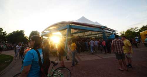 A lively outdoor event with a large tent, people mingling, and sunlight shining through the trees.