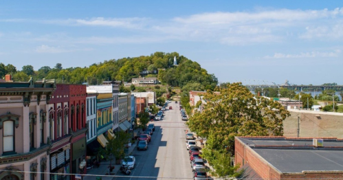 Aerial view of a small town street lined with colorful buildings, leading to a green hill with a statue on top.